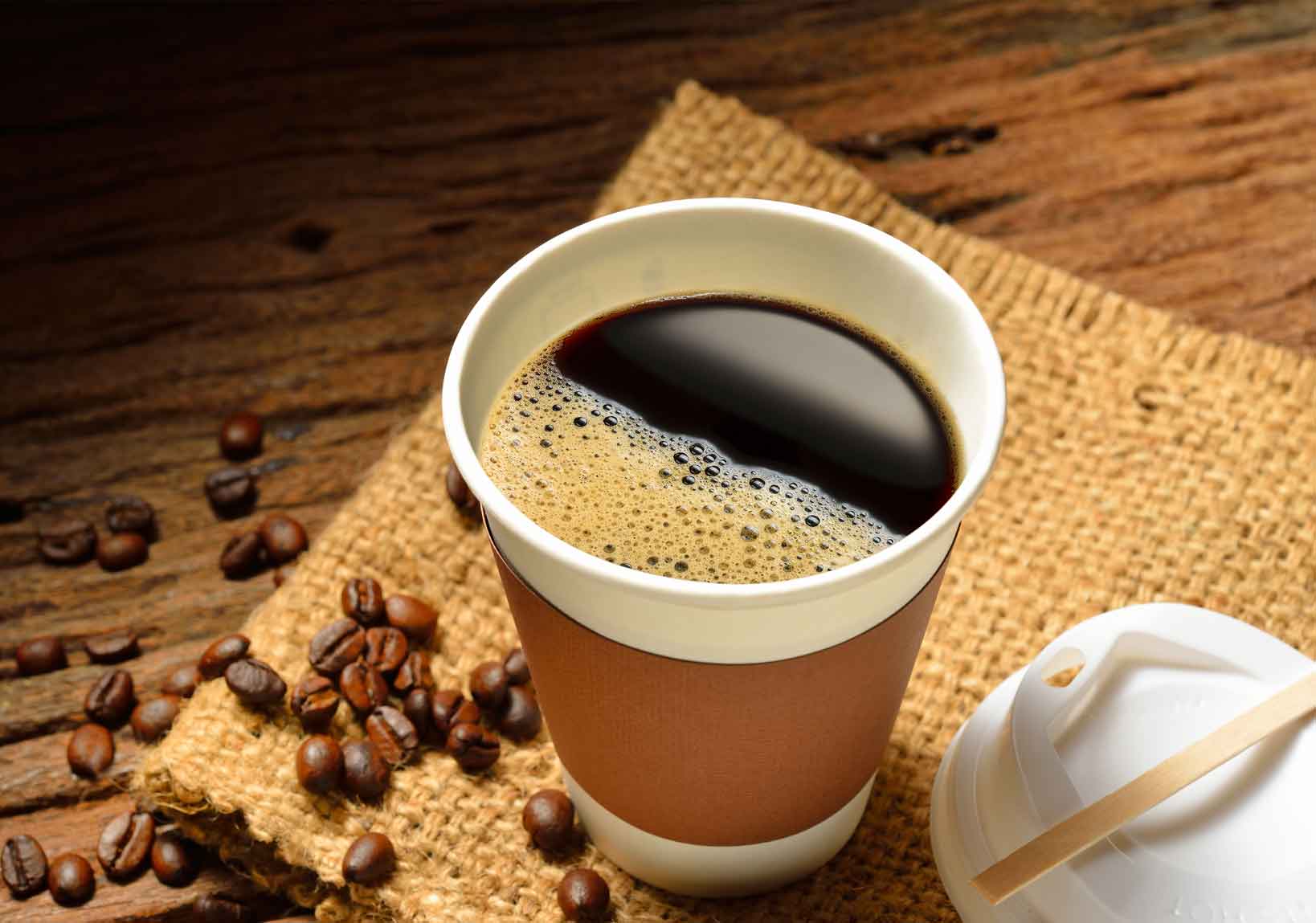 Paper cup of black coffee with foam on top, surrounded by scattered coffee beans on a burlap cloth and wooden table