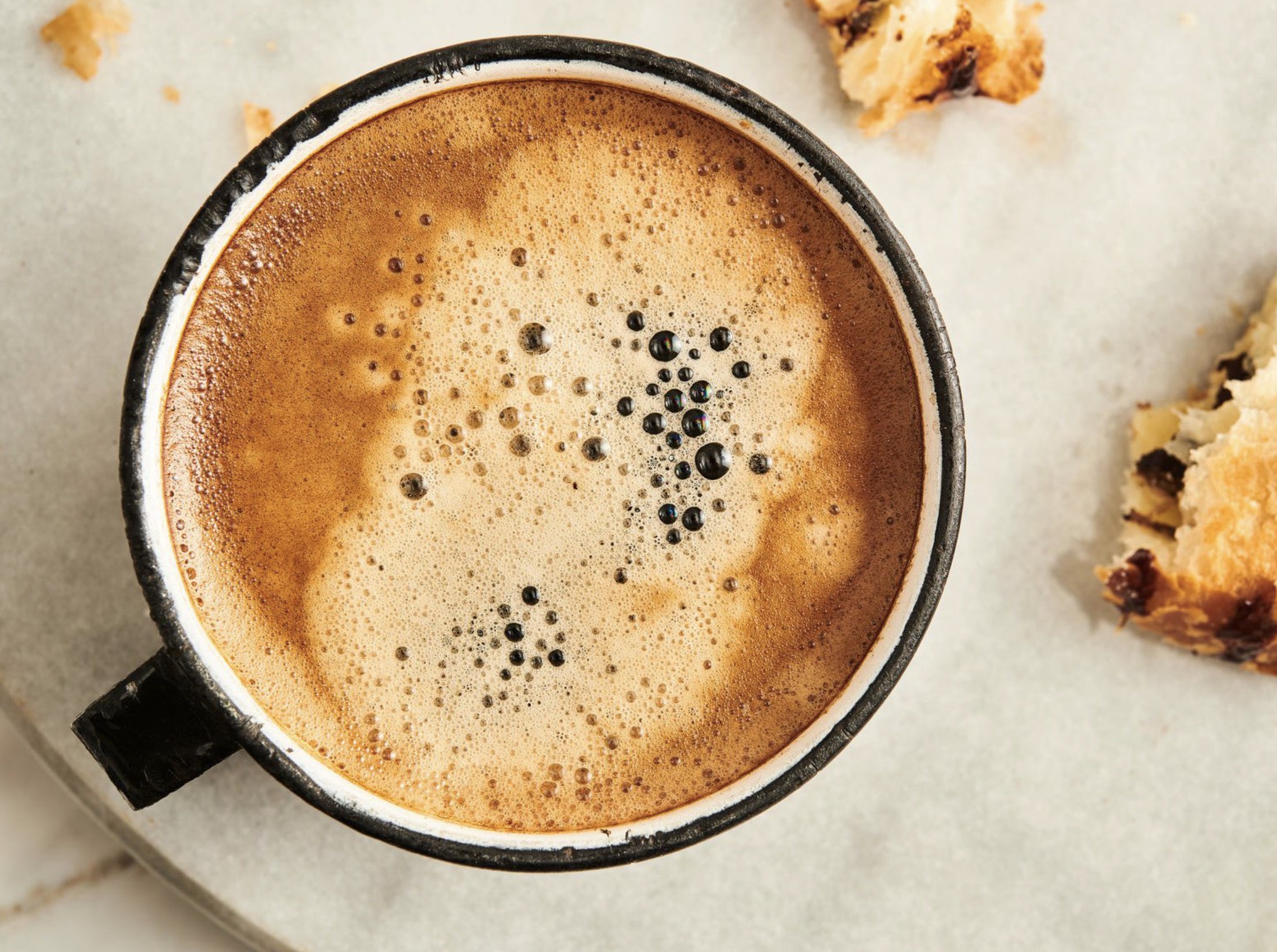 Overhead close-up of a frothy coffee in a dark rimmed mug with pastry crumbs beside it, warm brown and cream tones, cozy