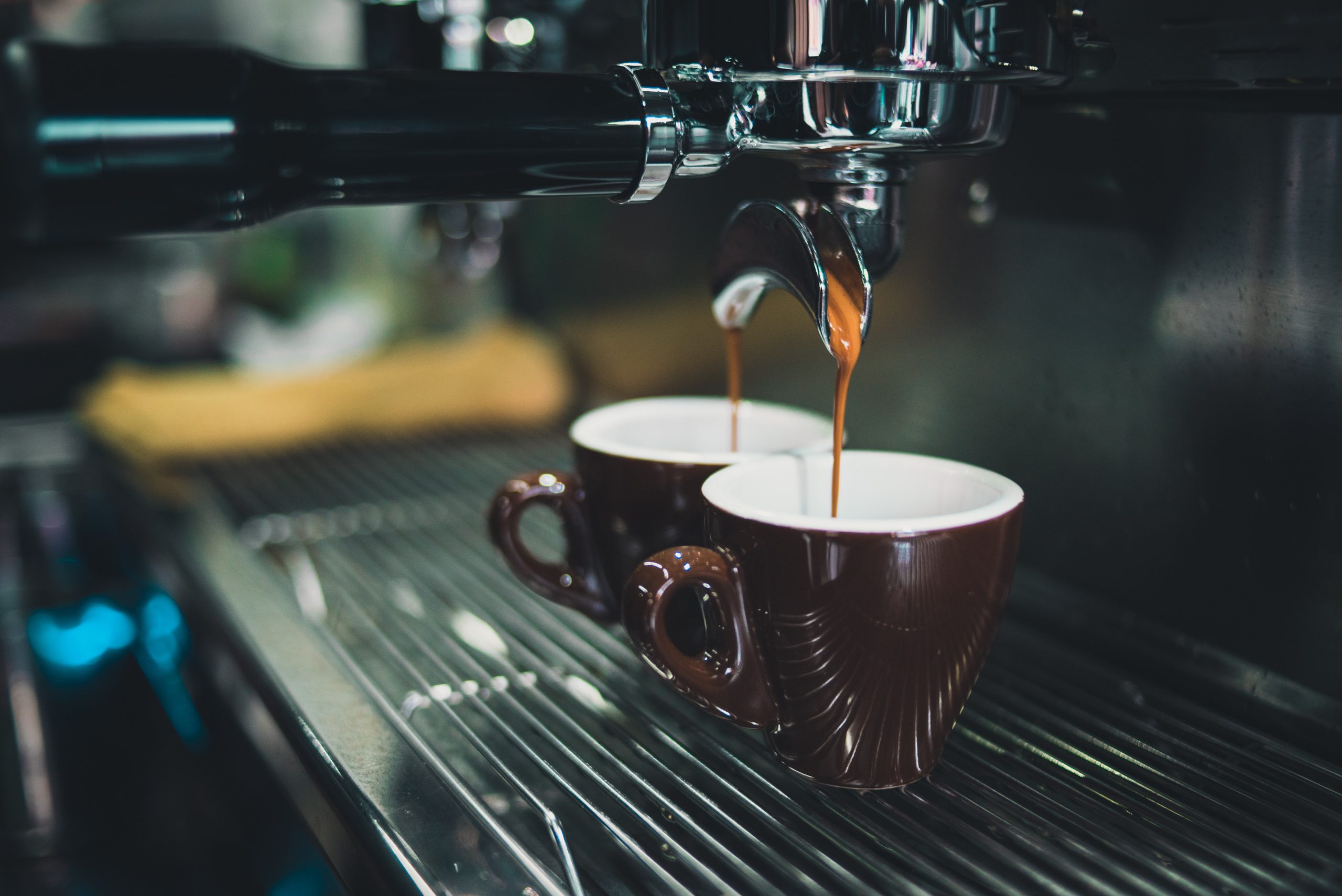 Close-up of an espresso machine portafilter pouring dark coffee into two brown and white cups, warm dim lighting, cozy atmosphere